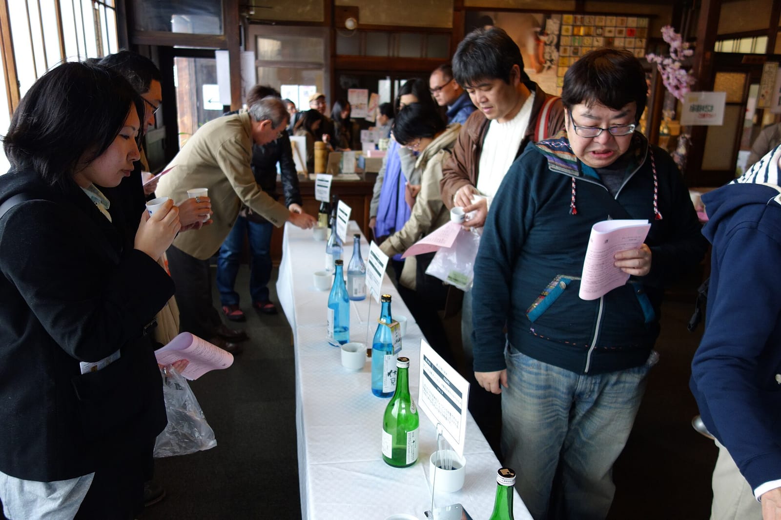 Adults holding small cups and printed tasting sheets beside a table lined with sake bottles at a crowded indoor tasting.