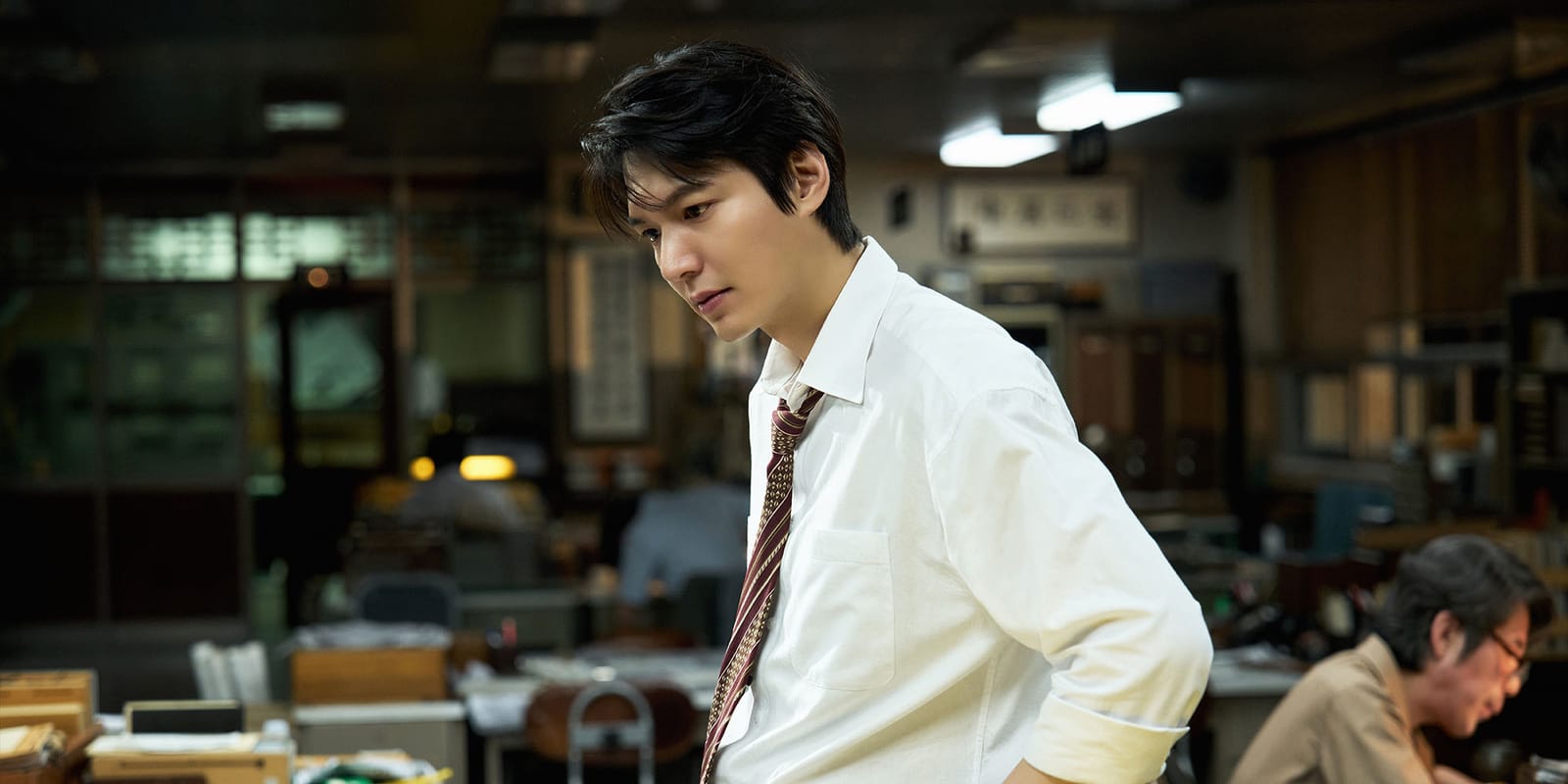 Film still from The Assassin(s) showing a man in a white shirt and loosened tie in a crowded office