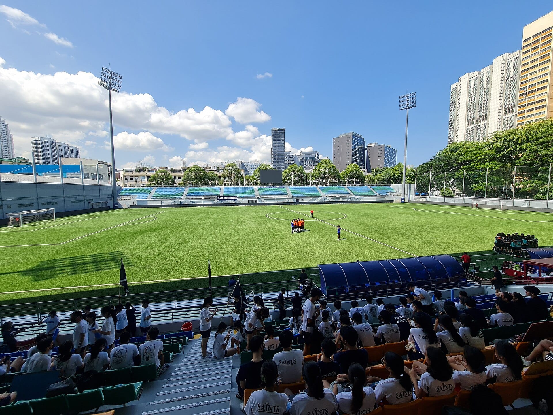 Football teams huddle on the artificial-turf pitch at Jalan Besar Stadium as spectators watch from the covered stand.